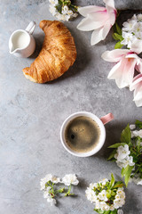 Obraz na płótnie Canvas Pink mug of black espresso coffee, french croissant, cream and spring flowers magnolia, blooming cherry branches over grey texture background. Top view, space. Spring greeting card