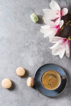 Blue cup of black espresso coffee, french dessert macaroons and spring flowers magnolia branches over grey texture background. Top view, space. Spring greeting card,
