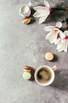 Pink mug of black espresso coffee french dessert macaroons, cream and spring flowers magnolia branches over grey texture background. Top view, space. Spring greeting card
