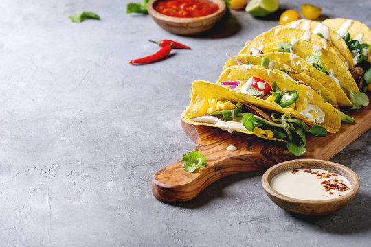 Variety of vegetarian corn tacos with vegetables, green salad, chili pepper served on olive wooden cutting board with tomato and cream sauces with ingredients above over grey texture background.