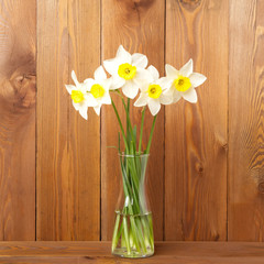 Bouquet of fresh flowers, daffodils in vase in middle of wooden table, opposite brown wooden wall. Empty space for text.