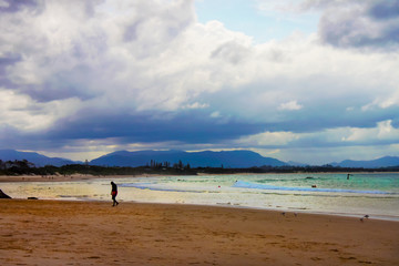 Man slumping - tired or sad - walking on beach coming out of surf at golden hour with beach full of people and mountians and stormy sky in the background