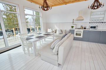 Wide angle shot of modern open living room interior overlooking floor to ceiling windows with white wooden table and grey kitchen counters in minimalistic Scandinavian design by sunlight, copy space