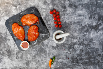 Raw marinated chicken breast fillets on black stone plate tray with BBQ sauce in white dish on grey concrete background, prepatation for grilling or smoking, top view, overhead