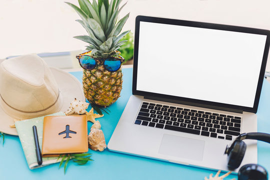 planning summer vacation concept. stylish laptop with empty screen, pineapple in sunglasses and passport, map, hat, shells, notebook on trendy blue paper table. travel and wanderlust