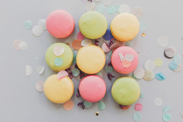 Fototapeta na wymiar delicious colorful macaroons on trendy pastel gray paper with lilac flowers and confetti, flat lay. tasty pink, yellow, green macaroons. candy for party. yummy image for instagram