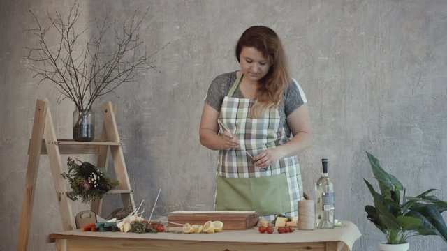 Concentrated attractive redhead woman in apron pinning slices of assorted types of cheese on wooden skewers in workshop while preparing edible bouquet composition for a gift.