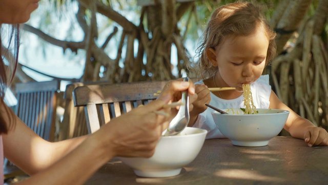 Mother and daughter eats noodles soup via chopsticks at the open air beach cafe - Powered by Adobe