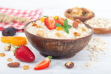 Oatmeal porridge in bowl with fresh strawberries, nuts and dried fruit. Decorated with mint leaves. Wooden spoon, top view