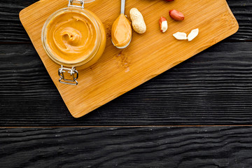 Make peanut butter. Paste in glass jar on cutting desk near peanut on black wooden background top view copy space