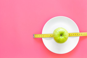 Proper nutrition with dietary fibre for weight loss. Apple on plate near measuring tape on pink background top view copy space