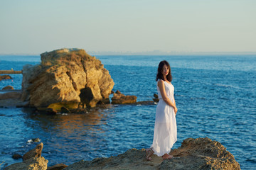 Fototapeta na wymiar Young beatiful woman wearing white dress standing on rock in blue sea. Looking on amazing sideview, horizone and smiling. Feeling good, free, happy. Summer, warm, good weather.