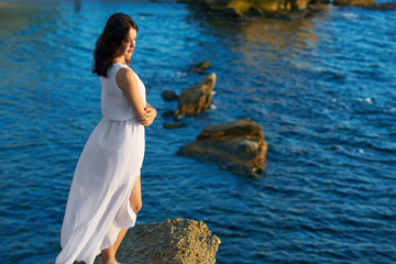 Backview of young beatiful model wearing white dress standing on high rock in blue sea. Looking on amazing sideview, horizon and smiling. Feeling good, free, happy. Summer, warm, good weather.