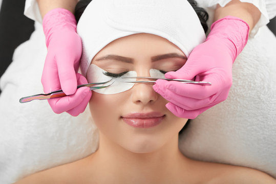Photo of lashmaking in modern beaty salon. Professional is making long lashes for a client wearing pink hygienic gloves. Master is using different types of tweezers. Smiled model with closed eyes.