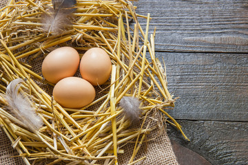 three chicken eggs lie in a nest of straw with feathers and down on a wooden table. view from above. place for text