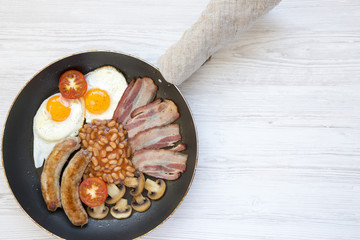 Full English Breakfast in cooking pan with sausages, mushrooms, fried eggs, beans, tomato and bacon on a white wooden background. Copy space. Top view. Flat lay. From above.