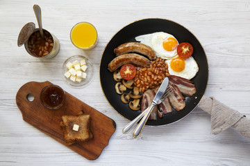 English breakfast in a frying pan with fried eggs, bacon, sausages, beans and toasts on white wooden background, top view. Flat lay. From above.