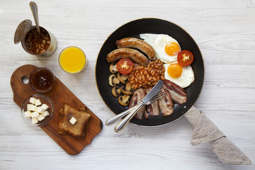 English breakfast in a frying pan with fried eggs, bacon, sausages, beans and toasts on white wooden background, top view. Flat lay. From above.