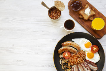 Full English breakfast in a pan with fried eggs, bacon, sausages, beans and toasts on white wooden background, top view. Copy space. Flat lay. From above.