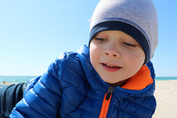 Cute boy in warm clothes lying on sandy beach on sunny day. 