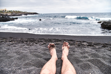 Men's legs on the beach. A unique beach with black volcanic sand. Waves splash about the shore, a fresh sea breeze is blowing. A man, a guy, a boy is resting on the beach.