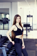 Fototapeta na wymiar Portrait of Young adult fit sporty girl poising near shelf with metal dumbbell in gym room space. She is looking right at camera. Attractive woman has slim body with press. Vertical photo