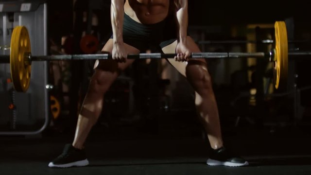 Tilt up of sexy muscular woman in sports bra and shorts picking up barbell from the floor in gym and doing squats