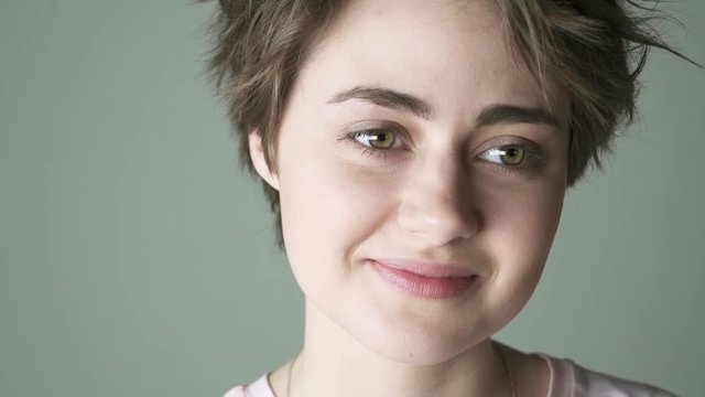 Beautiful teenage girl with short brown hair wearing a pink t shirt thinking while standing against a gray background. Handheld slow motion close up shot