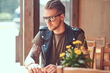 Hipster with a stylish haircut and beard sits at a table in a roadside cafe.