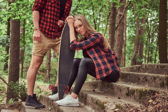 Young hipster couple, handsome man and blonde girl with a skateboard, sitting on steps in a park.