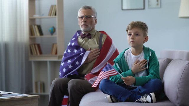Patriotic old man holding American flag, singing national anthem with grandson
