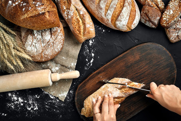 Woman hands cutting a loaf of bread on rustic wooden board, with wheat ears and knife, top view.