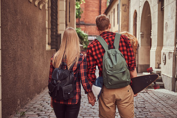 Back view of a young hipster couple, handsome skater and his girlfriend, holding hands, walking around a old narrow streets of Europe. - Powered by Adobe
