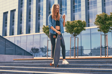 Young hipster blonde girl in casual clothes, standing on steps against a skyscraper, using a smartphone, resting after riding on the skateboard.