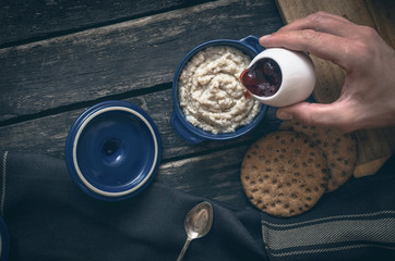 Barley porridge with strawberry jam in the blue ceramic pot and bread on dark aged wooden background. Healthy breakfast. Man pours porridge with jam.