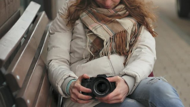 Young beautiful woman in a red hat wearing sporty warm clothes and rollers, sitting on a wooden bench and taking pictures on a vintage camera
