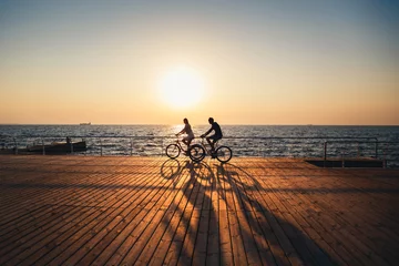 Schilderijen op glas Couple of young hipsters cycling together at the beach at sunrise sky at wooden deck summer time © Iryna Budanova