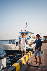 Cute young beautiful couple at pier at port with small yachts, hipster, happy smiling outdoor portrait