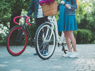 Naklejka premium View of young couple walking together with retro white and modern black bicycles in the city park. Basket with peony flowers. Romantic scene. Summer season. Vintage hipster color toned.