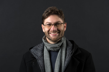 Bearded man in scarf and coat on dark background. Happy man in glasses on bearded face. Fashion model with beard smile on dark background. Autumn style and trend. Fashion accessory concept