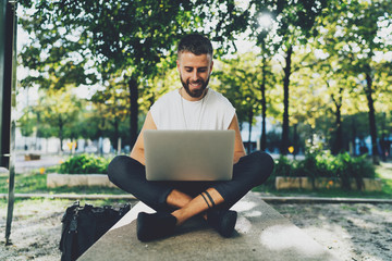 Fototapeta premium Portrait of young handsome bearded male typing on a laptop keyboard while sitting outdoors with a computer on his knees. Student guy in casual outfit surfing the web on a computer connected to wifi