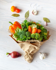 Bouquet of fresh vegetables lies on a white wooden table. Nearby is a red onion, tomatoes, garlic, bay leaf. View from above