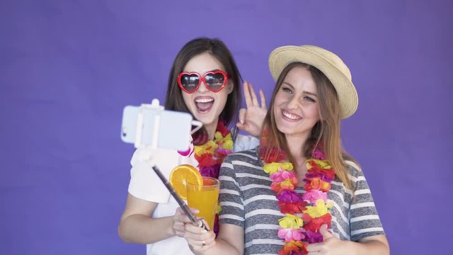 Delighted young women taking selfie with a selfie stick, wearing hawaiian wreaths around the slim necks, isolated shot in the purple background