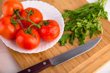 Fresh red tomatoes close up on a white plate at a wooden board with green parsley and a knife