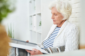 Side view portrait of elegant senior woman opening box of photos and cherished little things remembering her family and friends while sitting alone at home in modern apartment.