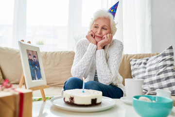 Portrait of lonely senior woman nervously celebrating birthday alone with photograph of her late husband on table by birthday cake photo in frame by me 