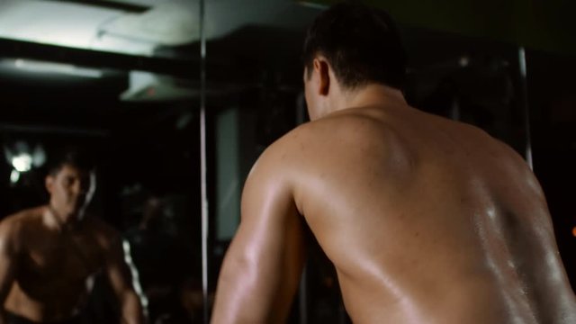 Shirtless muscular athlete doing cable rope lateral raise exercise in front of the mirror in dark gym