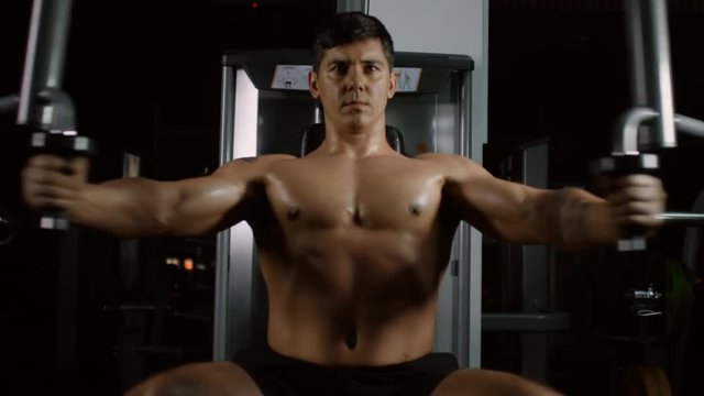 Muscular handsome man doing pec deck butterflies and looking at camera while exercising on fitness machine in gym