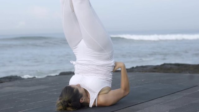 Medium shot of flexible mature woman in white doing shoulder stand on coastline near ocean