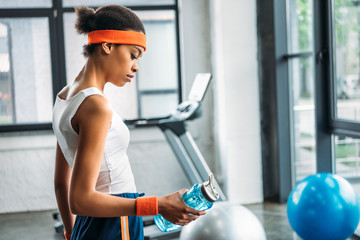 side view of african american sportswoman in headband and wristband holding bottle of water at gym